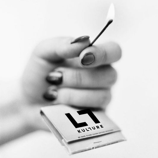A hand with painted nails holds a lit match above an open KULTURE "LT" Matches 5Pk by Kulture, Inc., the box labeled "KULTURE" in bold black text and capturing the unique allure of this collectible.